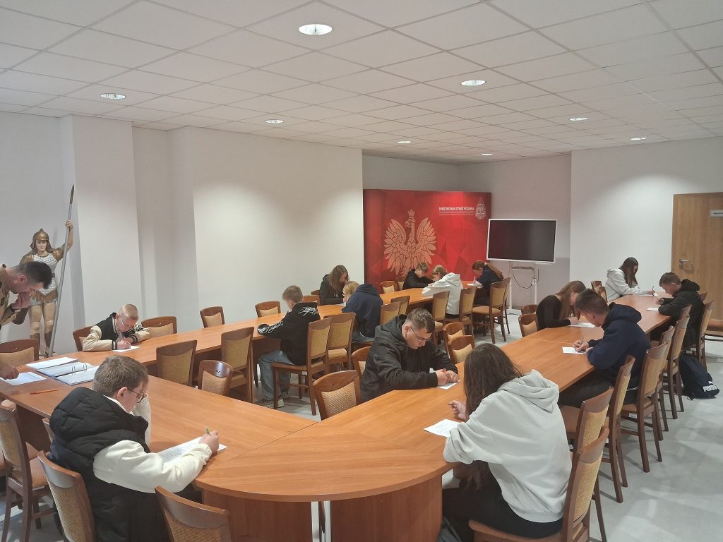 Grupa młodych osób siedzi przy drewnianych stołach w sali z białymi ścianami, pisze na kartkach, obok stoi figurka wojownika z włócznią.