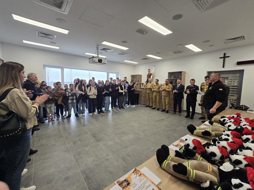 Grupa dzieci i dorosłych stoi na tle białych ścian w jasnym pomieszczeniu, po prawej stronie stoją mężczyźni w mundurach i garniturach, na pierwszym planie pluszowe misie w strażackich ubraniach.