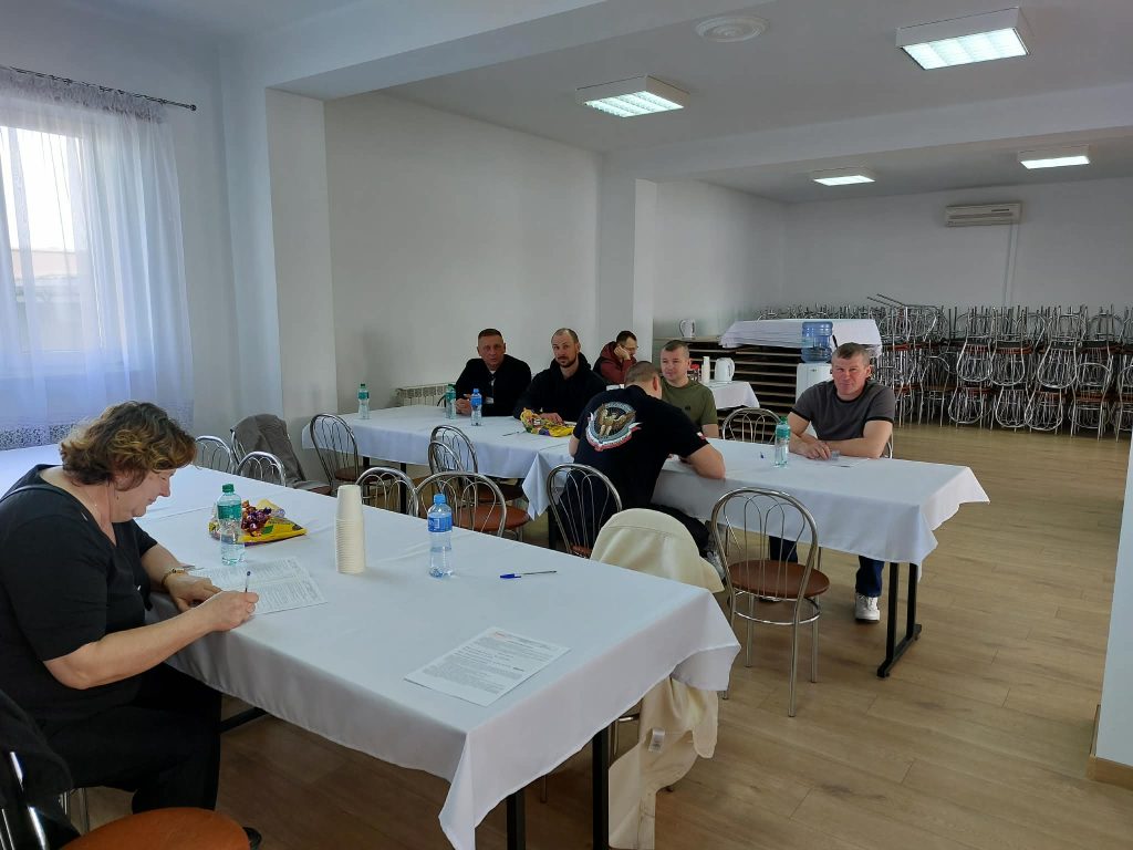 Kilkoro dorosłych siedzi przy białych stołach z metalowymi krzesłami w jasnym pomieszczeniu z drewnianą podłogą i dużym oknem z białą firanką.