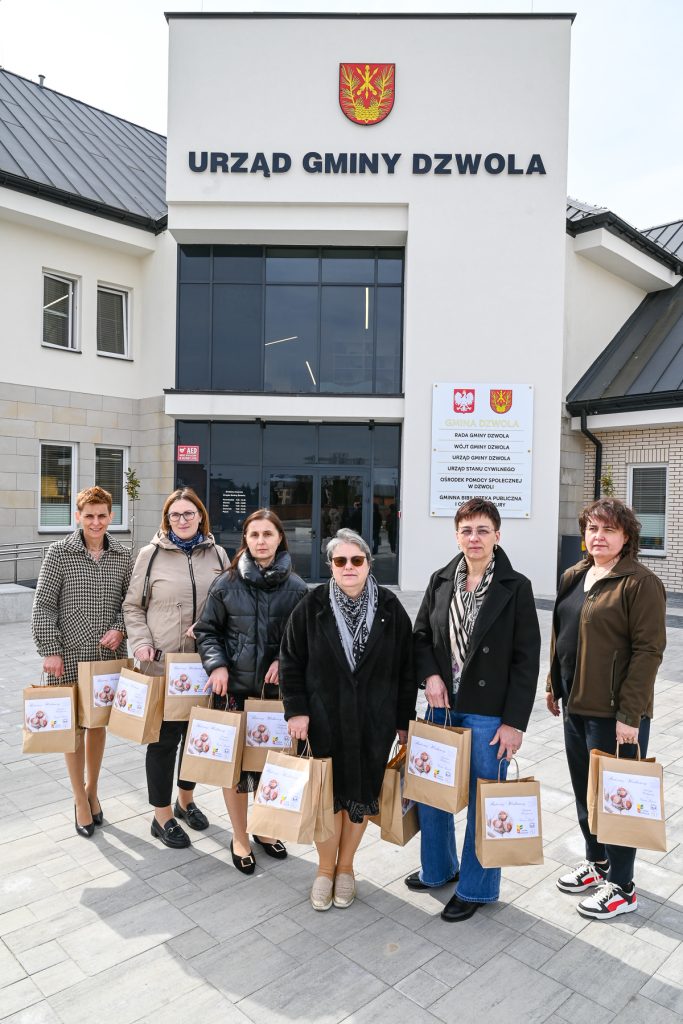 Sześć kobiet stoi przed budynkiem Urzędu Gminy Dzwola, każda trzyma brązową torbę z białą etykietą, ubrane w różne płaszcze i okulary przeciwsłoneczne.
