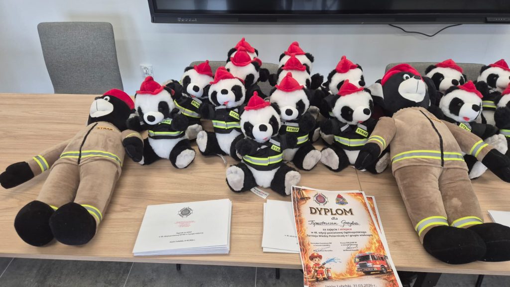 Pluszowe misie panda w czarnych strażackich kurtkach i czerwonych czapkach, pluszowe misie w brązowych strażackich ubraniach, dyplomy i dokumenty na drewnianym stole.