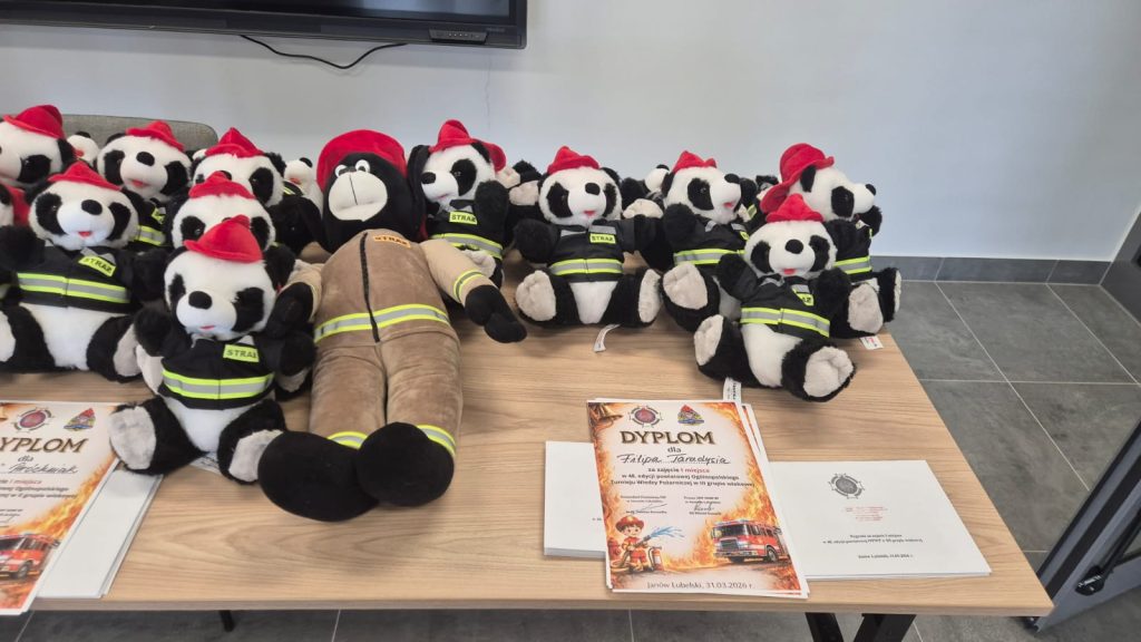 Pluszowe misie panda i niedźwiedź w czarnych kurtkach strażackich i czerwonych hełmach na jasnym stole obok dyplomów z ilustracją wozu strażackiego.