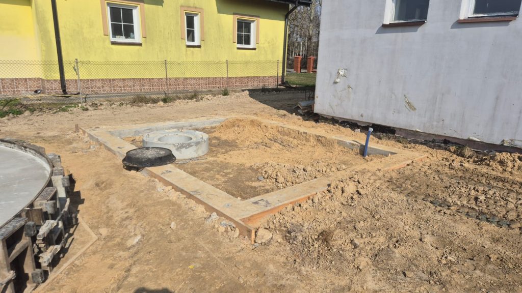 Plac budowy z piaszczystym podłożem, fundamentem z betonu i rurą wystającą z ziemi, obok żółty budynek z oknami i ogrodzenie z siatki metalowej.