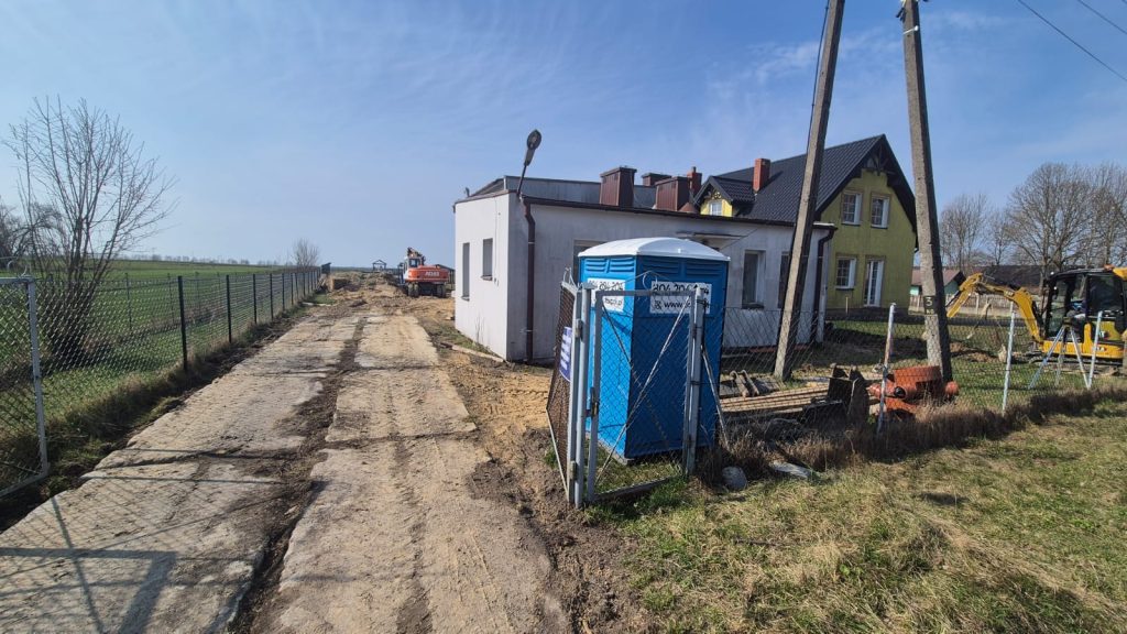 Niebieska toaleta przenośna obok białego budynku z ogrodzeniem, przydrożna droga z betonowych płyt, koparka i maszyny budowlane na tle zielonych pól i niebieskiego nieba.