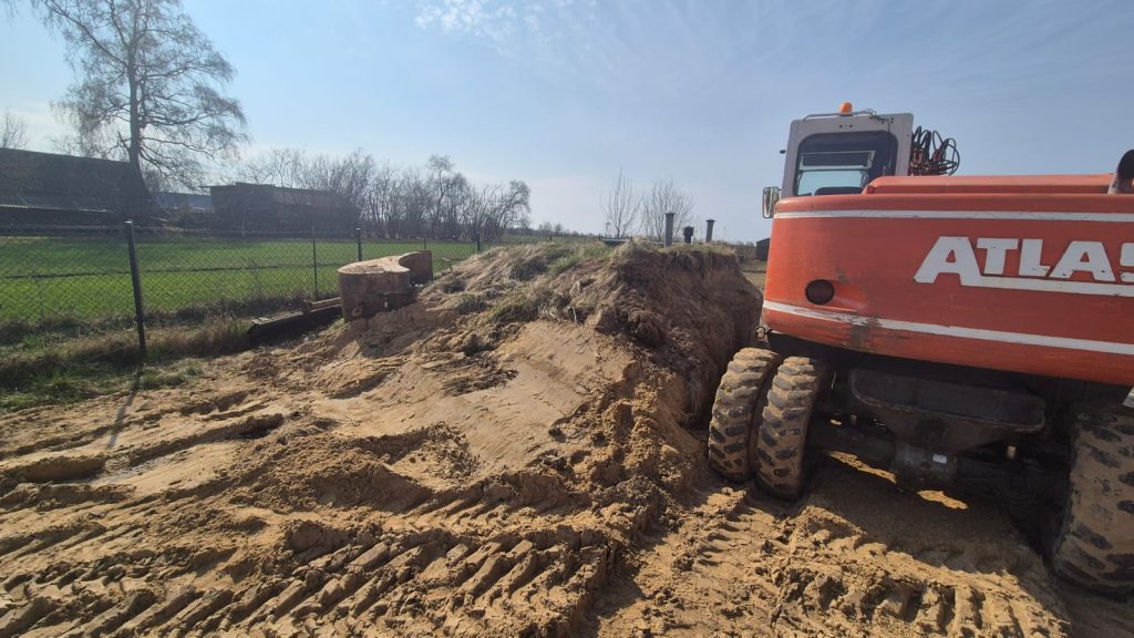 Czerwony sprzęt budowlany na piaszczystym placu z wyraźnymi śladami opon, trawiasta ziemia i metalowy zbiornik obok, w tle zielona łąka i drzewa.