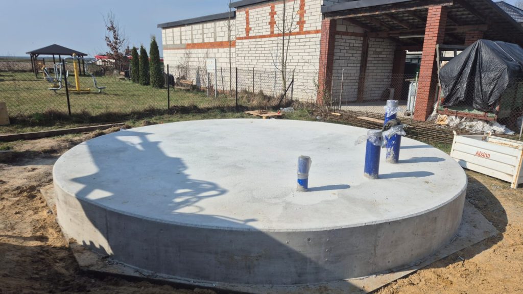 Okrągła betonowa płyta z trzema pionowymi niebieskimi rurami na placu budowy, w tle ogrodzenie, budynek i urządzenia do ćwiczeń na świeżym powietrzu.
