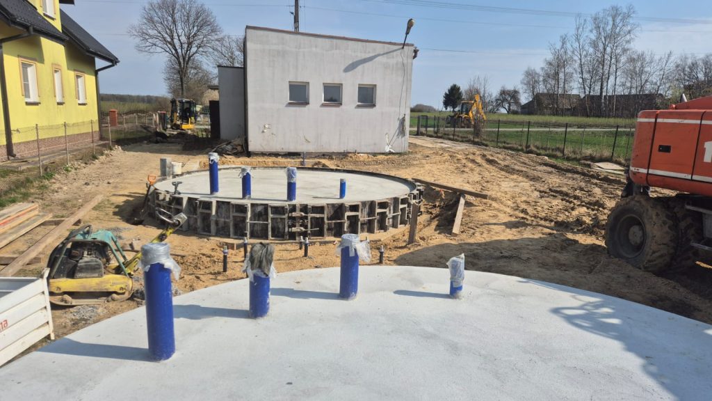 Plac budowy z dwoma okrągłymi betonowymi fundamentami z niebieskimi rurami, obok maszyny budowlane i żółty budynek.