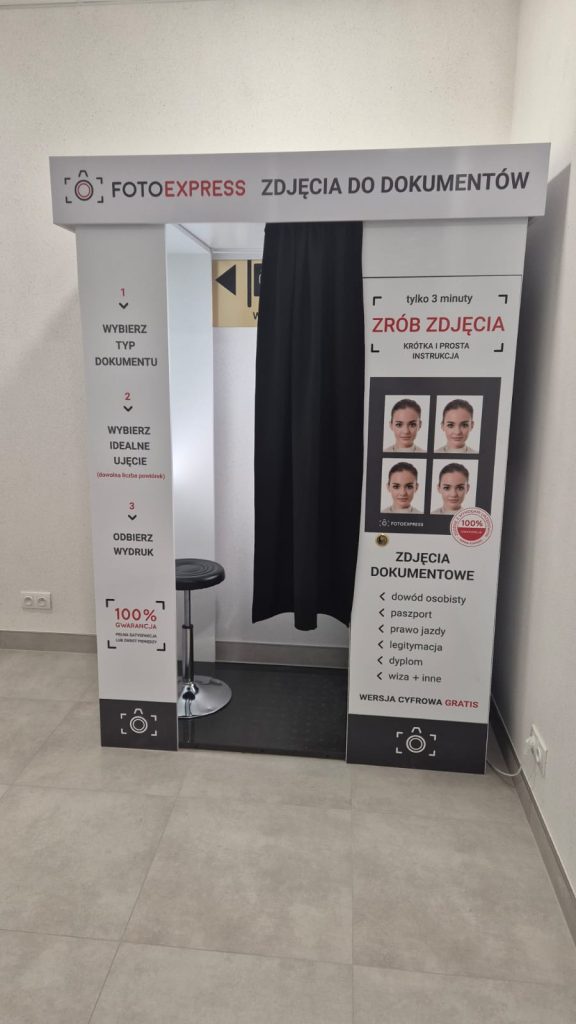 Biała budka do zdjęć dokumentowych z czarną zasłoną, tablicą z instrukcjami i zdjęciami przykładowymi, obok metalowy stołek, jasna podłoga i ściana.