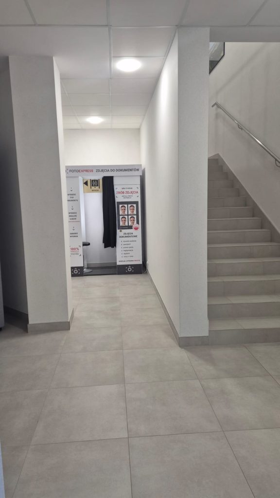 Wnętrze z jasnoszarymi płytkami podłogowymi, białymi ścianami, schodami po prawej stronie i kabiną do zdjęć do dokumentów na końcu korytarza.