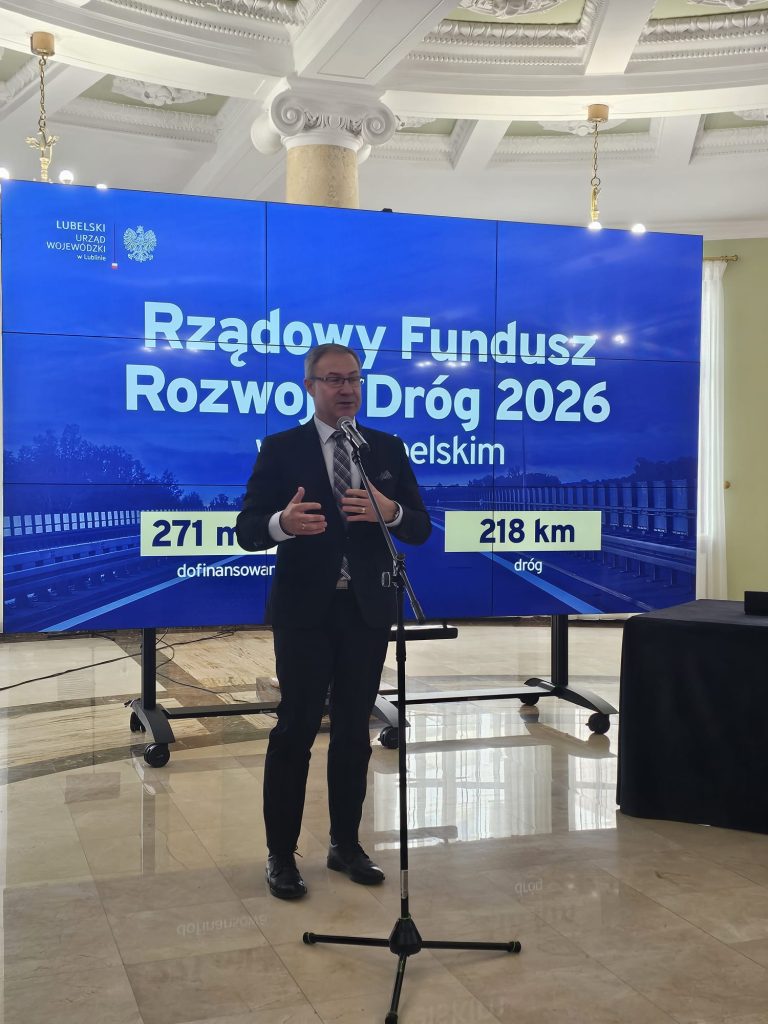 Mężczyzna w garniturze przemawia przy mikrofonie na tle niebieskiego ekranu z napisem o rządowym funduszu rozwoju dróg 2026, marmurowa podłoga i kolumna.
