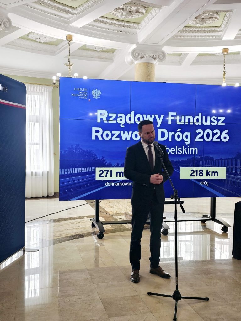 Mężczyzna w ciemnym garniturze stoi przy mikrofonie na tle dużego ekranu z niebieskim tłem i białym napisem oraz kolumną i jasną podłogą z marmuru.