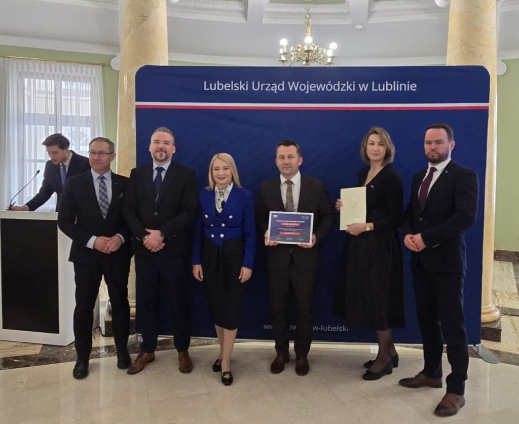 Sześć osób w formalnych ubraniach na tle granatowego baneru z napisem „Lubelski Urząd Wojewódzki w Lublinie”, dwie osoby trzymają dokumenty, sala z marmurową podłogą i żyrandolem.