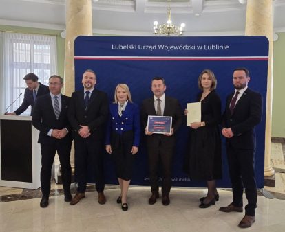 Sześć osób w formalnych ubraniach na tle granatowego baneru z napisem „Lubelski Urząd Wojewódzki w Lublinie”, dwie osoby trzymają dokumenty, sala z marmurową podłogą i żyrandolem.