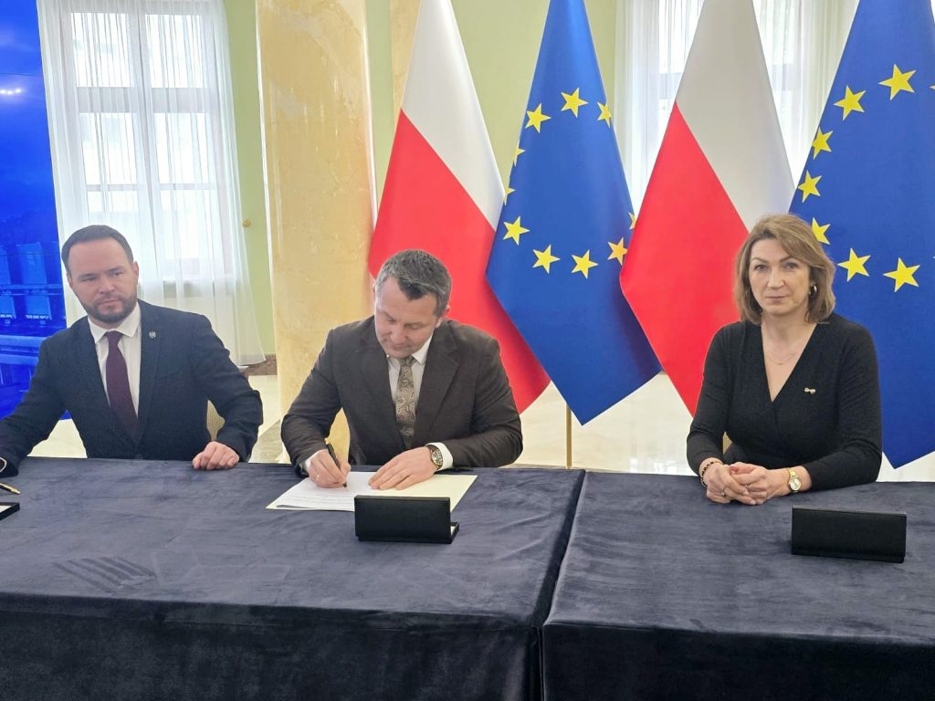 Trzy osoby w garniturach i eleganckiej odzieży siedzą przy ciemnym stole, za nimi flagi Polski i Unii Europejskiej na jasnym tle z oknami.