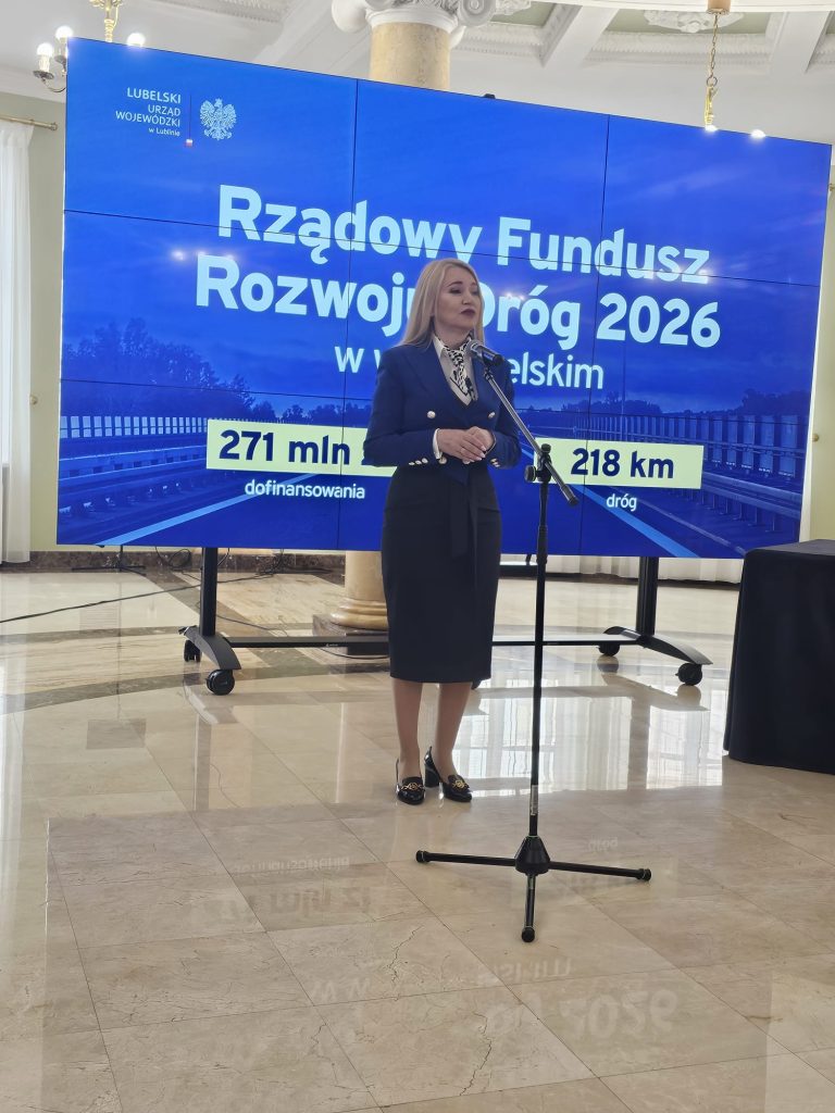 Kobieta w granatowym kostiumie stoi przy mikrofonie przed dużym niebieskim ekranem z napisem o rządowym funduszu rozwoju dróg 2026 i danymi finansowymi.