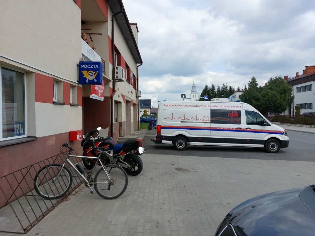 Biały van z napisem Regionalne Centrum Krwiodawstwa zaparkowany obok budynku poczty, przed wejściem stoją rower i czarno-czerwony motocykl, pochmurne niebo.