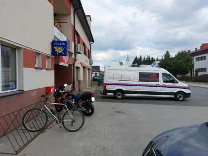 Biały van z napisem Regionalne Centrum Krwiodawstwa zaparkowany obok budynku poczty, przed wejściem stoją rower i czarno-czerwony motocykl, pochmurne niebo.