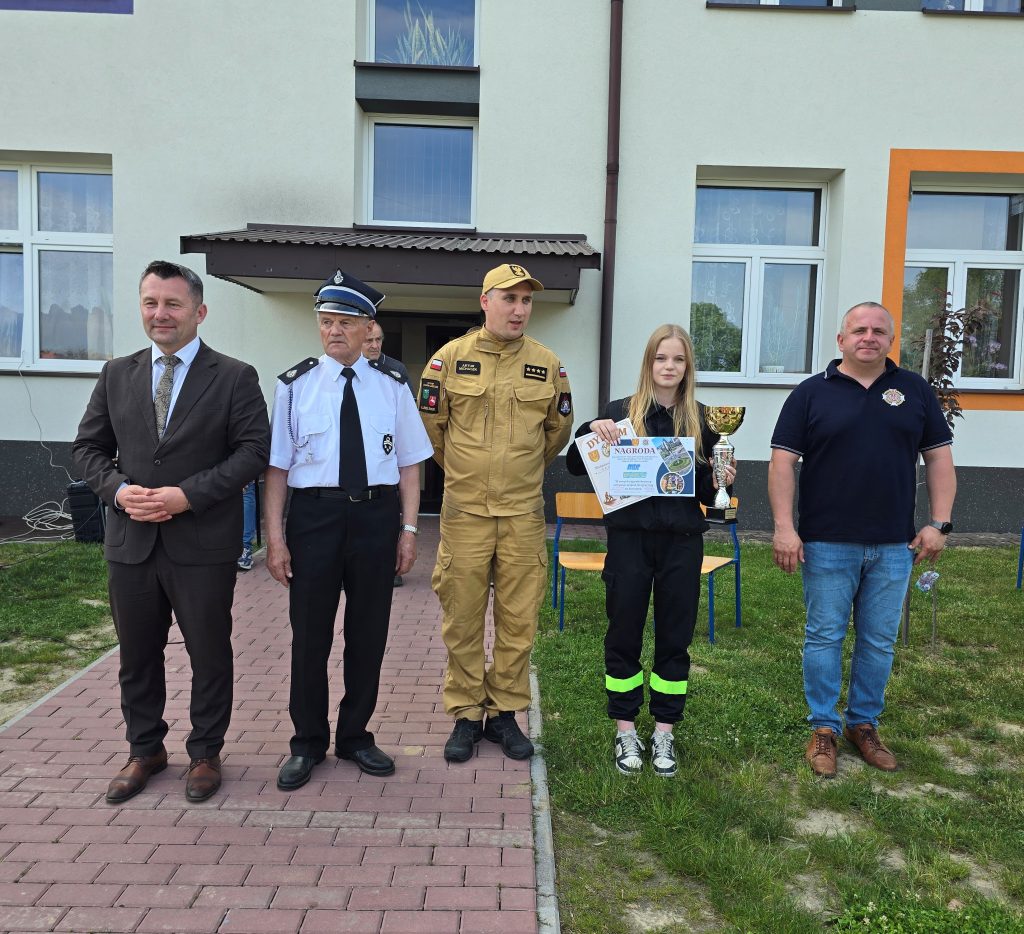 Grupa pięciu osób stojących na tle jasnej ściany budynku z oknami, jedna trzyma puchar i dyplomy, dwie osoby w mundurach, pozostałe w cywilnych ubraniach.