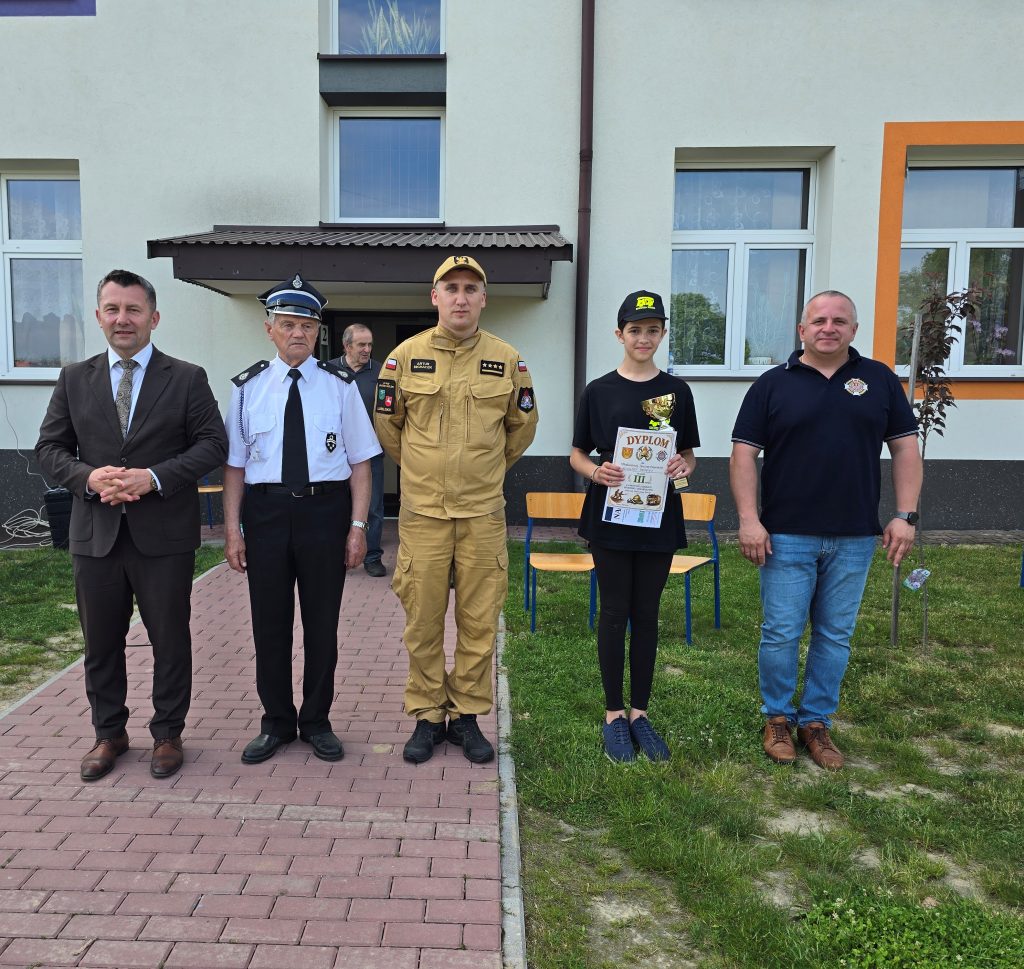 Pięć osób stojących na tle budynku, różne mundury i stroje, młoda dziewczyna z czarną czapką trzyma dyplom i puchar, trawnik i chodnik z czerwonej kostki brukowej.