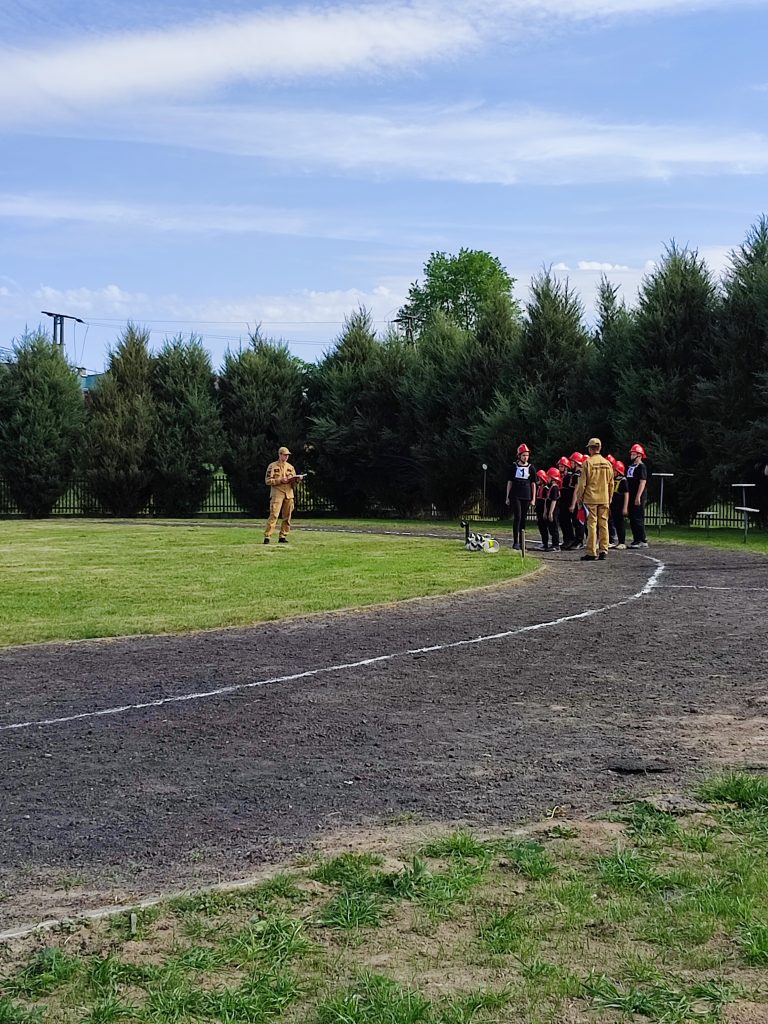 Grupa osób w czerwonych kaskach i czarnych ubraniach na torze z ziemi i trawy, otoczona wysokimi zielonymi drzewami, pod niebieskim niebem z chmurami.