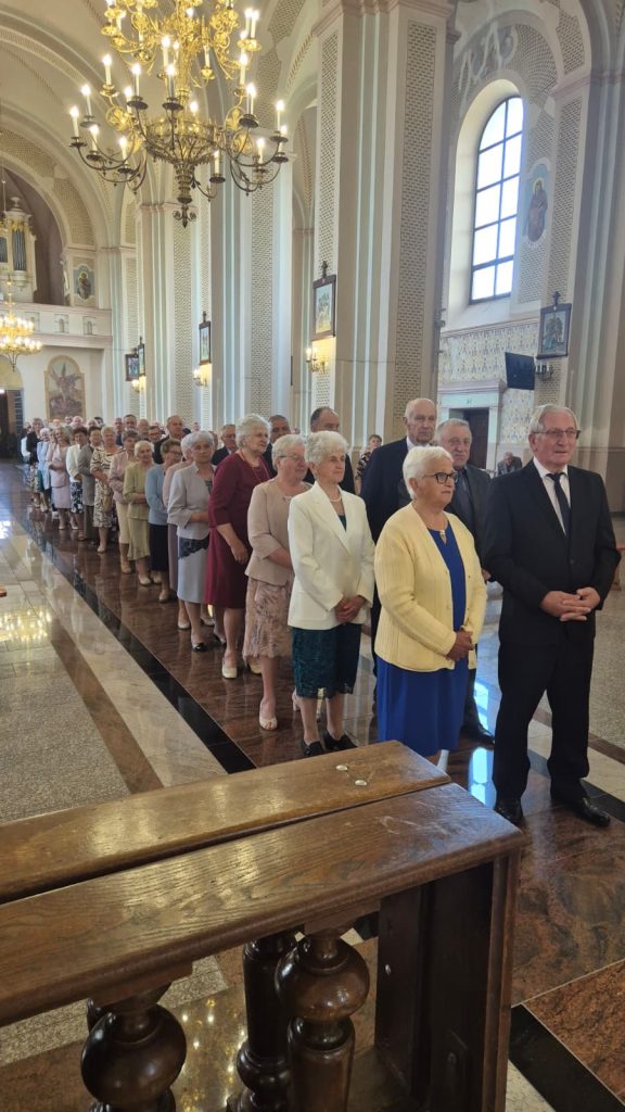 Kolejka starszych osób ubranych elegancko stojących w jasnym wnętrzu kościoła z dużymi oknami i złotymi żyrandolami.