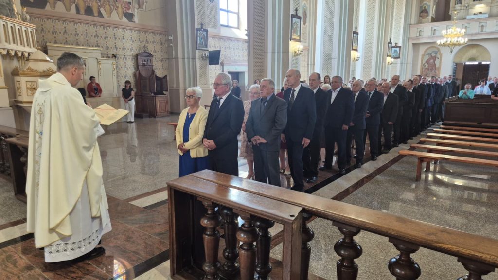Kolejka osób w eleganckich ubraniach stojąca w kościele przed księdzem w białej szacie, jasne wnętrze z obrazami i drewnianymi ławkami.