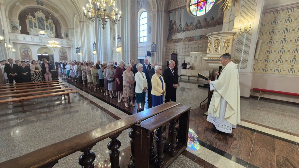 Wnętrze jasnego kościoła z wysokimi łukowatymi oknami, zdobieniami, witrażem i obrazem na ścianie, rząd starszych osób stojących przed duchownym w białej szacie.