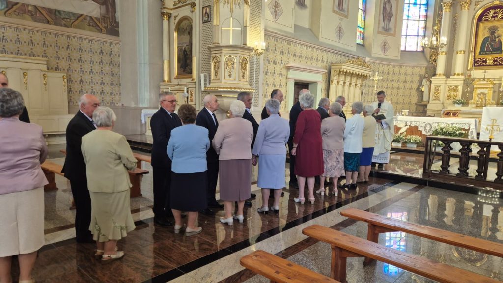 Grupa starszych osób w eleganckich strojach stoi w kolejce w jasnym, ozdobnym wnętrzu kościoła przed kapłanem przy ołtarzu.
