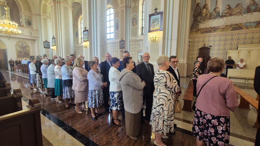Grupa starszych osób stojących w kolejce w jasnym, wysokim wnętrzu kościoła z obrazami i dużymi oknami.