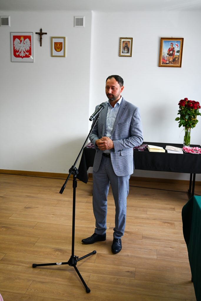 Mężczyzna w jasnoszarym garniturze stoi przy mikrofonie na stojaku na tle białej ściany z obrazami i krzyżem, obok stoi stół z bukietem czerwonych róż i dokumentami.