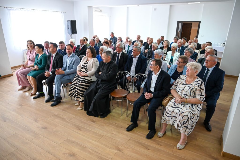 Grupa osób w różnym wieku ubranych formalnie, siedzących na metalowych krzesłach w jasnym pomieszczeniu z drewnianą podłogą i dużymi oknami.
