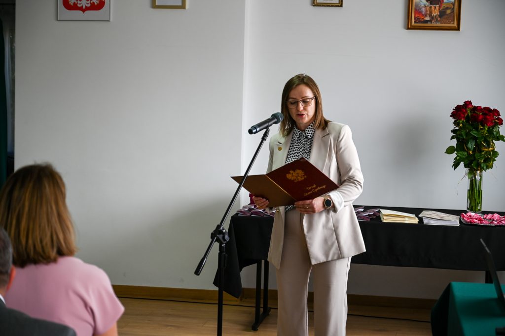 Kobieta w jasnym garniturze i okularach czyta z czerwonego folderu przy mikrofonie, obok stół z bukietem czerwonych róż, dokumentami i upominkami.