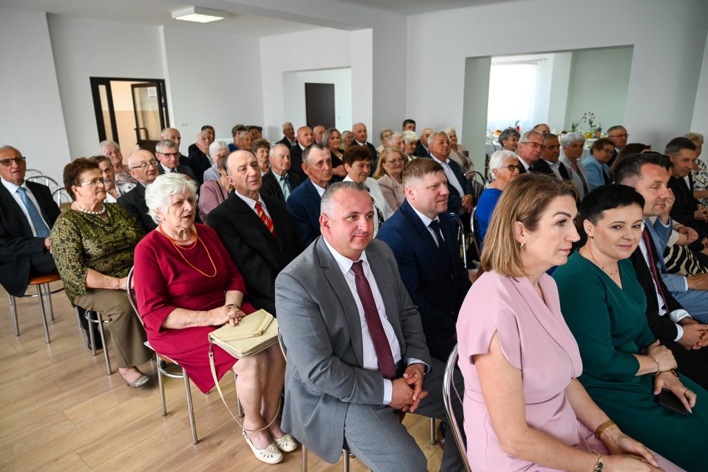 Grupa ludzi siedzących na metalowych krzesłach w jasnym pomieszczeniu, większość w formalnych strojach, kobieta w bordowej sukience z beżową torebką na pierwszym planie.