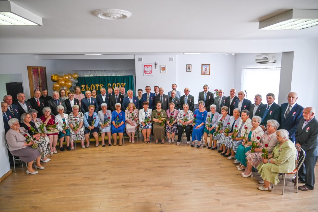 Grupa starszych osób w eleganckich ubraniach, kobiety siedzą z czerwonymi różami, mężczyźni stoją, w tle zielony baner z żółtym napisem i balony.