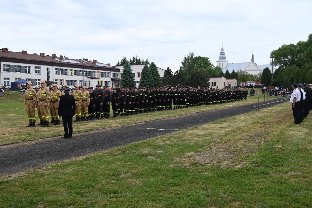 Linia osób w mundurach strażackich w dwóch kolorach, beżowym i czarnym, stojąca na trawiastym boisku, w tle budynki, drzewa i kościół.