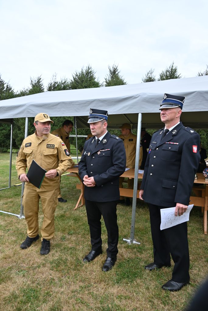 Trzech mężczyzn w mundurach, dwóch w granatowych z czapkami i jeden w beżowym z daszkiem, stoi na trawie przed namiotem, w tle zielone drzewa.