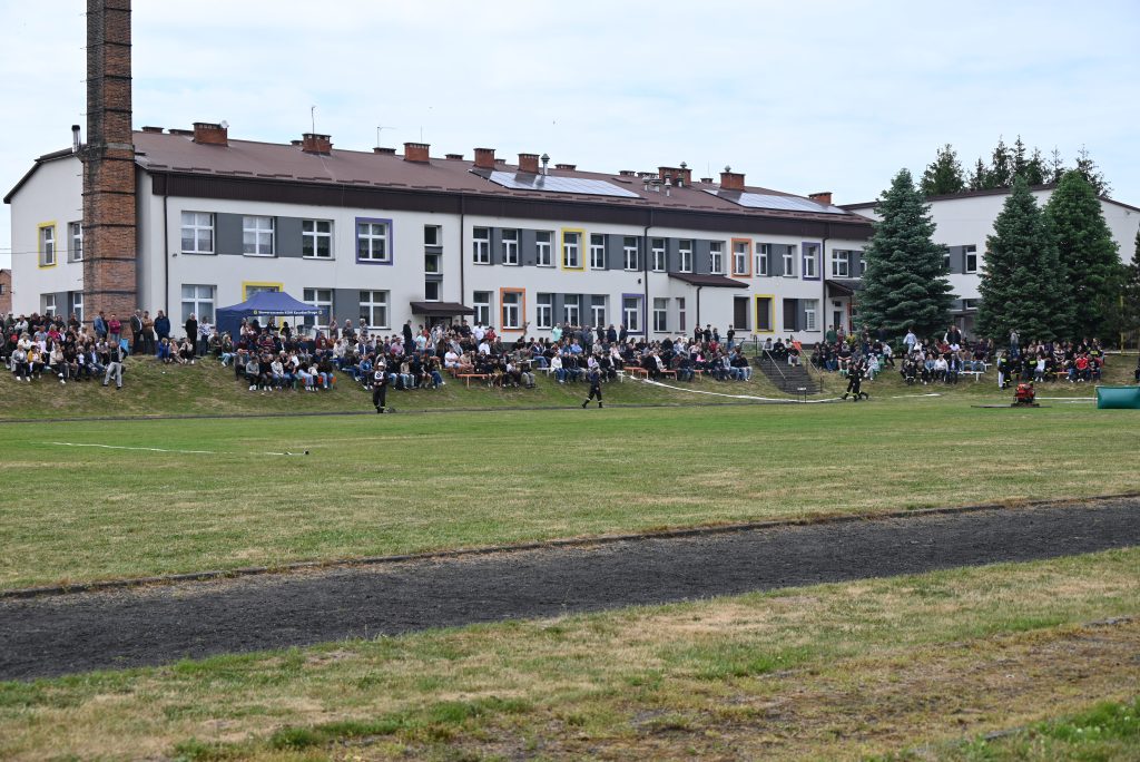 Biały budynek z kolorowymi okiennicami, przed nim trawiasta łąka i duża grupa ludzi siedzących na ławkach i stojących, obok wysokie choinki i komin z cegieł.