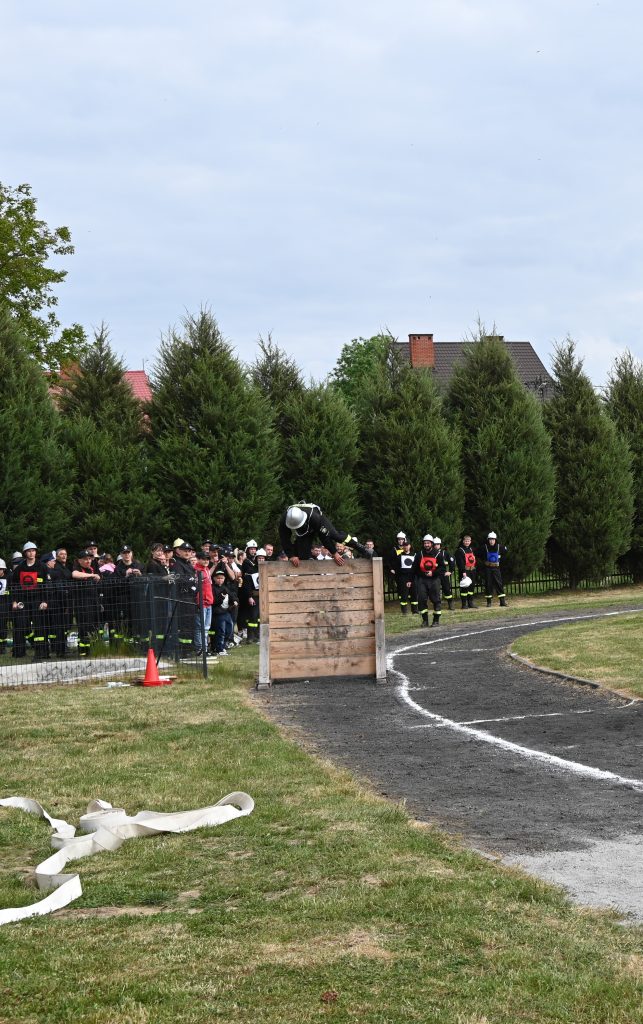 Grupa osób ubranych w czarne mundury i hełmy obserwuje uczestnika pokonującego drewnianą przeszkodę na trawiastym i szutrowym terenie.
