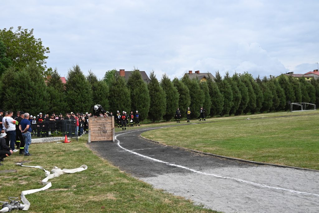 Grupa ludzi w mundurach strażackich na zielonym boisku, jeden skacze przez drewnianą przeszkodę, w tle drzewa i domy pod pochmurnym niebem.