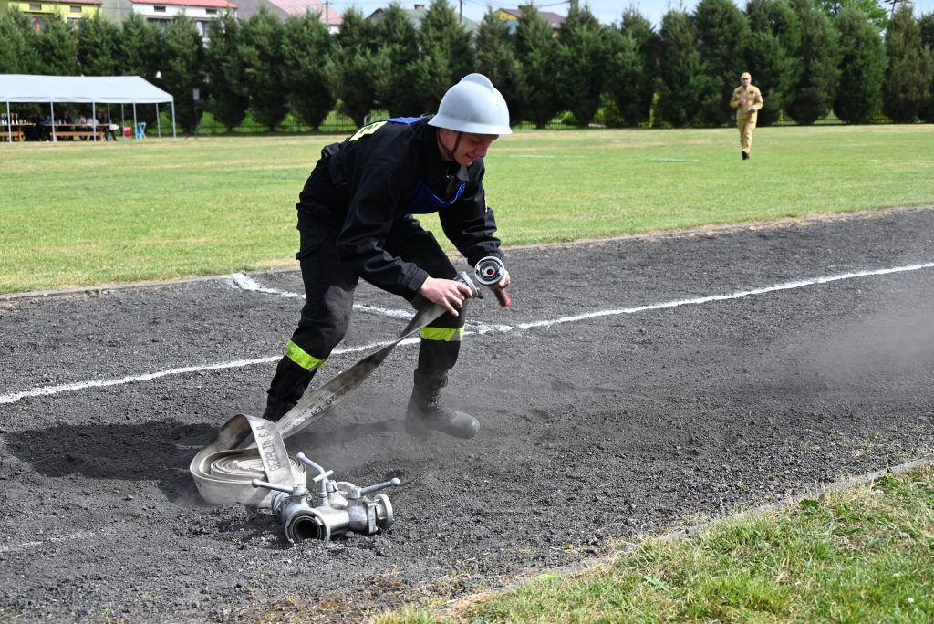 Strażak w czarnym ubraniu i białym hełmie rozwija beżowy wąż strażacki na czarnym podłożu obok metalowego rozdzielacza, w tle zielona trawa i drzewa.