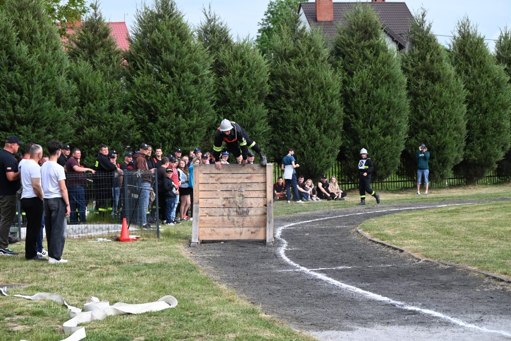 Mężczyzna w hełmie strażackim pokonuje drewnianą przeszkodę na bieżni, obok tłum obserwujących i zielone drzewa na tle domów.