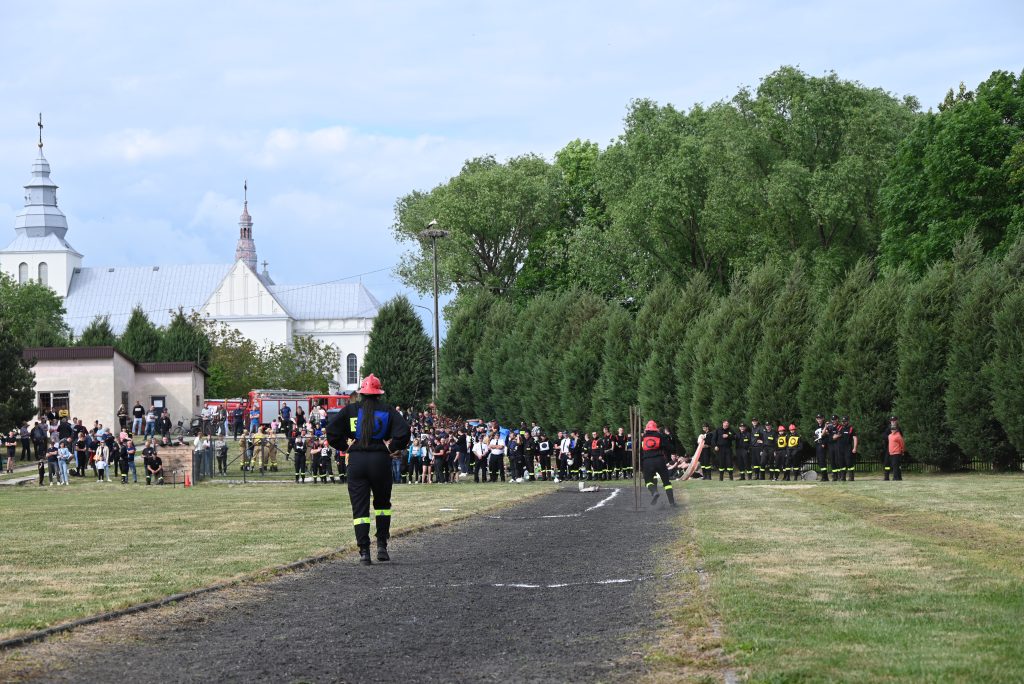 Duża grupa ludzi na trawie i na ścieżce, dwie osoby w kombinezonach strażackich i czerwonych hełmach, w tle biała budowla z wieżą i zielone drzewa.