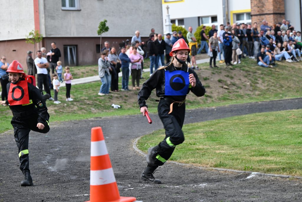 Dwóch młodych strażaków w czerwonych hełmach i czarnych mundurach biegnie po szutrowej ścieżce z pomarańczowym pachołkiem, w tle grupa ludzi na trawie i chodniku.