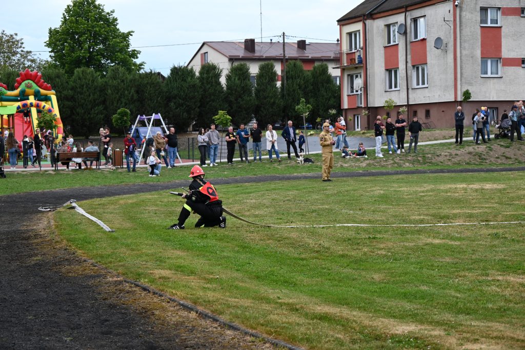 Osoba w stroju strażaka z hełmem i wężem strażackim na trawie, obok stoi mężczyzna w beżowym mundurze, w tle ludzie na trawniku i plac zabaw z dmuchaną zjeżdżalnią.