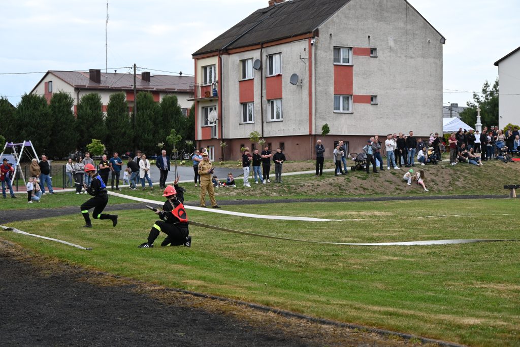 Dwóch strażaków w czarnych mundurach i czerwonych hełmach ćwiczy na zielonym trawniku, wokół nich stoi grupa ludzi, w tle szare budynki z czerwonymi elementami.