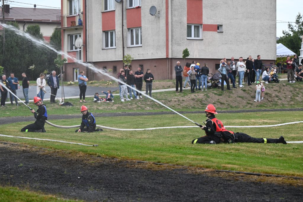 Cztery osoby w strojach strażackich i czerwonych hełmach trzymają węże z wodą, w tle ludzie stojący na trawie przed budynkiem mieszkalnym.