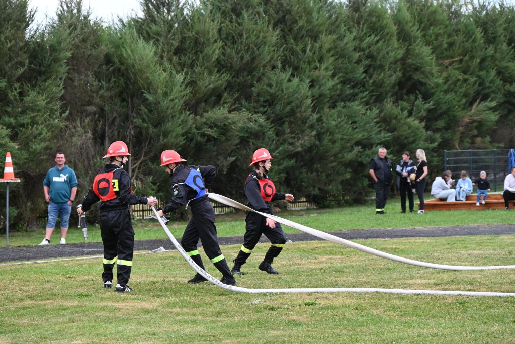 Trzech młodych strażaków w czarnych uniformach z czerwonymi kaskami na zielonym trawniku, w tle ludzie, drzewa i pachołek drogowy.