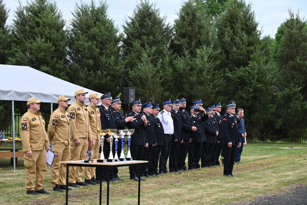 Grupa osób w mundurach beżowych i czarnych ustawionych w rzędzie na trawie, przed stołem z pucharami, w tle zielone drzewa i biały namiot.