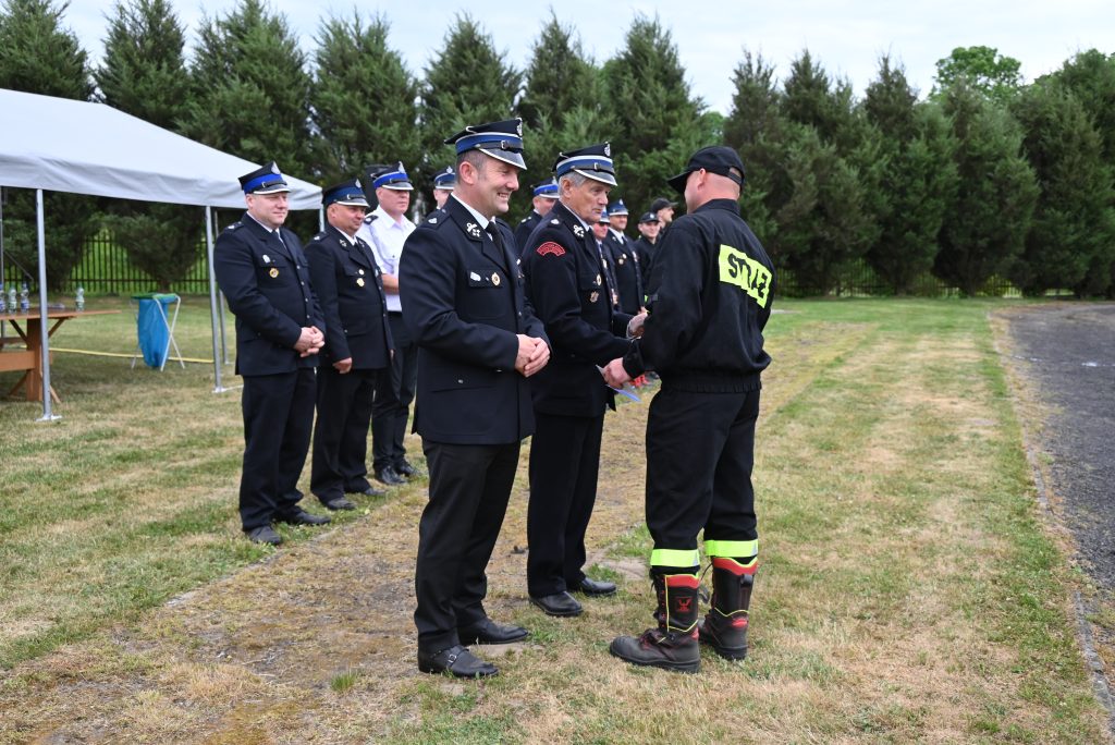Grupa mężczyzn w mundurach strażackich stoi na trawie obok namiotu, jeden z nich w czarnej odzieży z napisem "STRAŻ" na plecach. Tło to zielone drzewa.