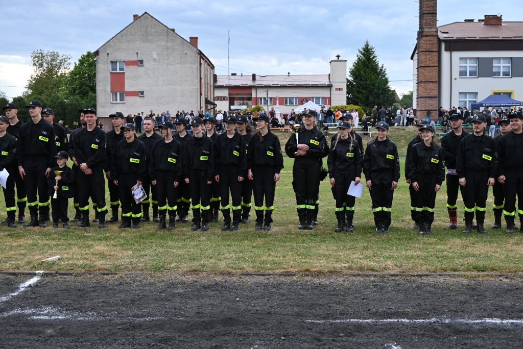 Grupa osób w czarnych mundurach z żółtymi napisami "STRAŻ", stojących na trawie przed budynkami, w tle tłum ludzi i niebo z chmurami.